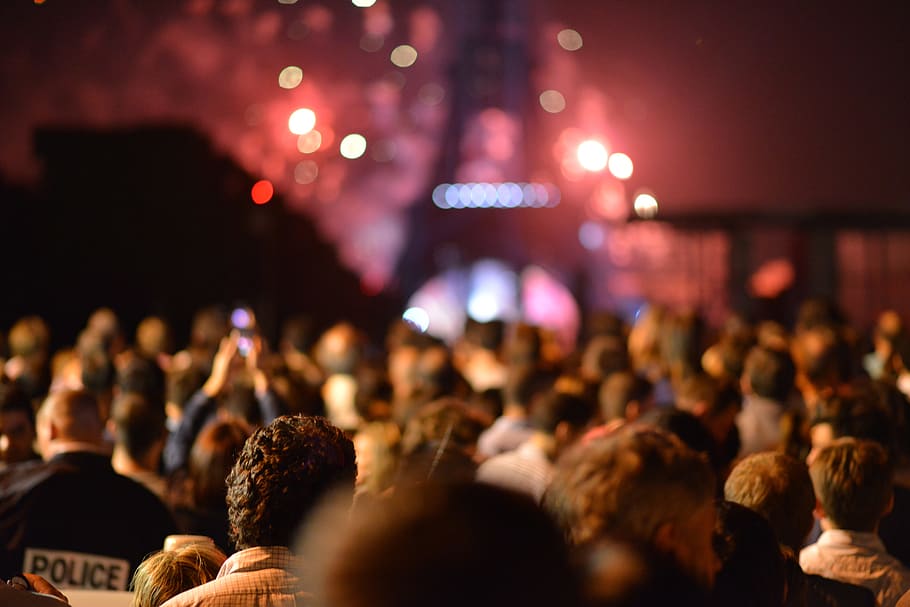 This photo displays a crowd of people who stand in front of the Eiffel Tower and who are watching the red colors of fireworks which seems to be happening behind and around the Eiffel Tower. On the lower left side a policeman is visible. HD wallpaper