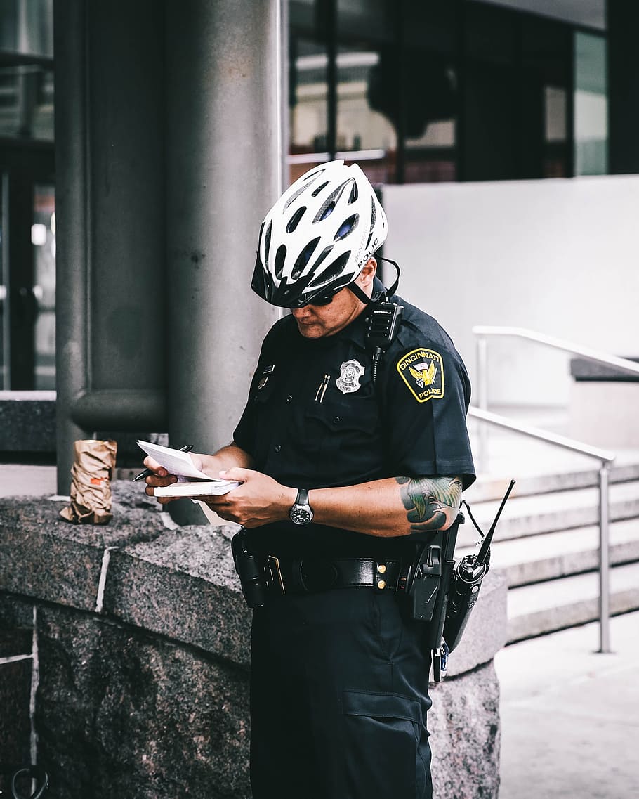 officer reading notes, Policeman holding white notepad and black pen standing near gray concrete bench outside building