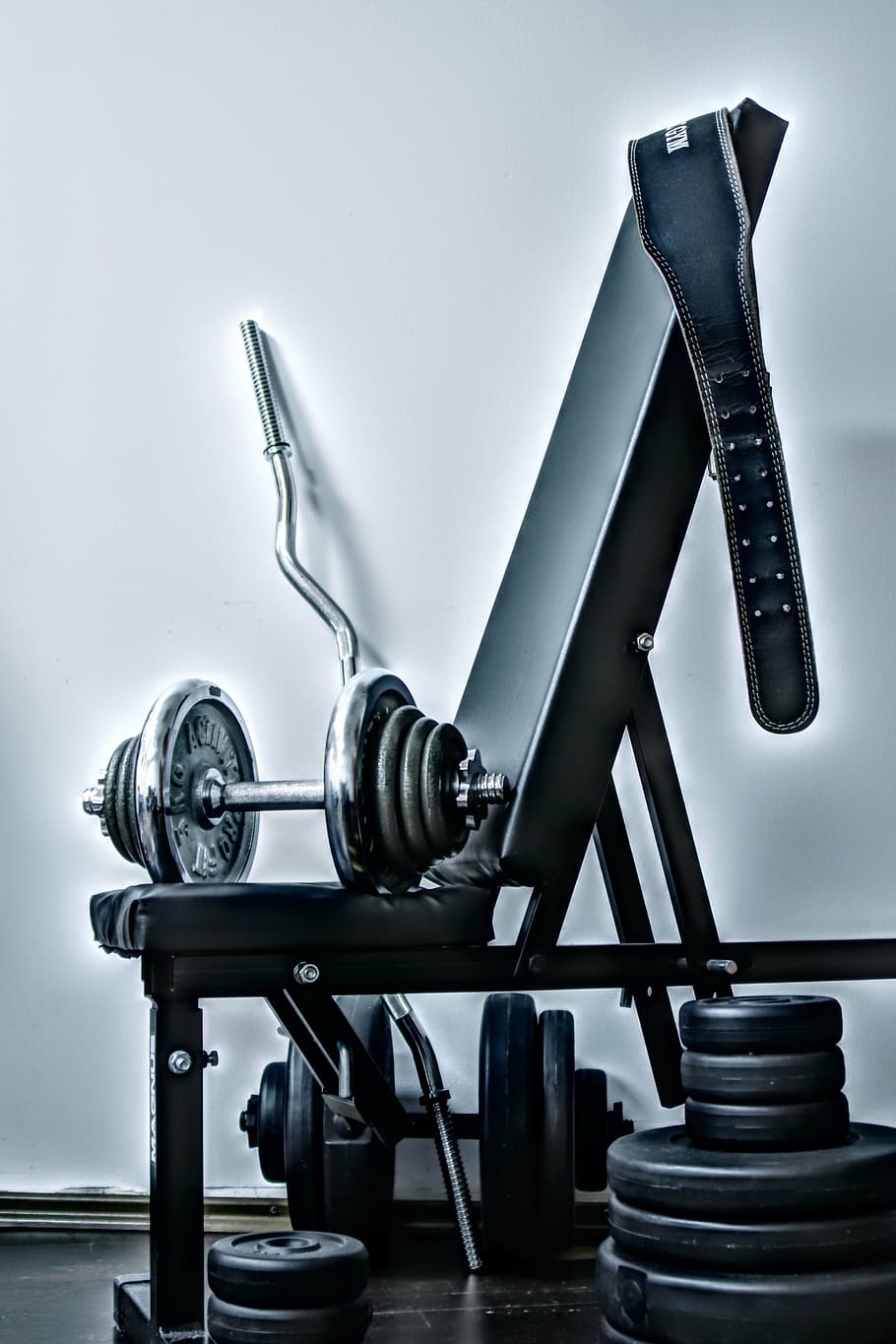 gray barbell and blue gym belt placed on black and gray exercise bench near wall
