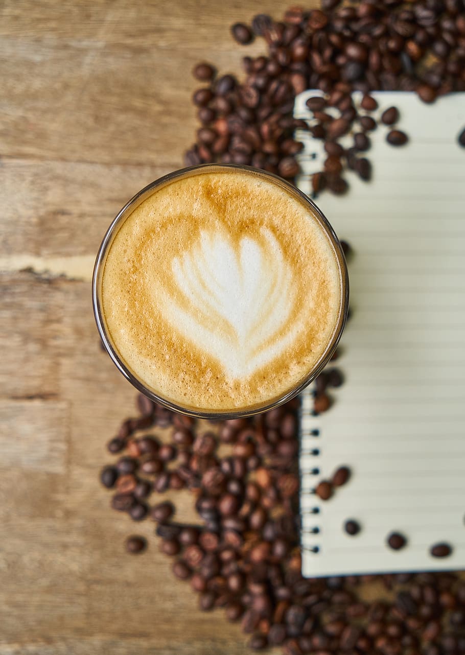 latte surrounded by coffee beans near white notepad on brown wooden surface HD wallpaper
