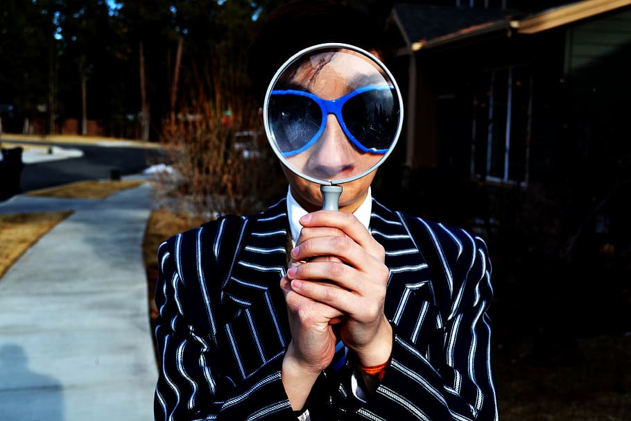 person using magnifying glass enlarging the appearance of his nose and sunglasses, man holding magnifying glass near green wooden house