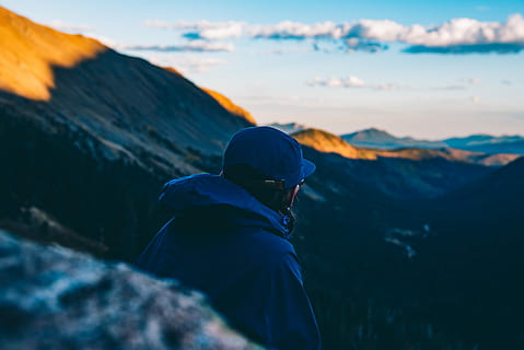 person wearing blue hoodie and blue fitted cap sitting near cliff looking at mountain range under white clouds during daytime, man sitting looking at mountain HD wallpaper