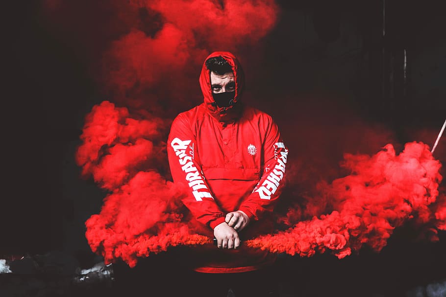 man wearing red and white hoodie and black mask surrounded by red smoke, man in red Thrasher hoodie holding red spray