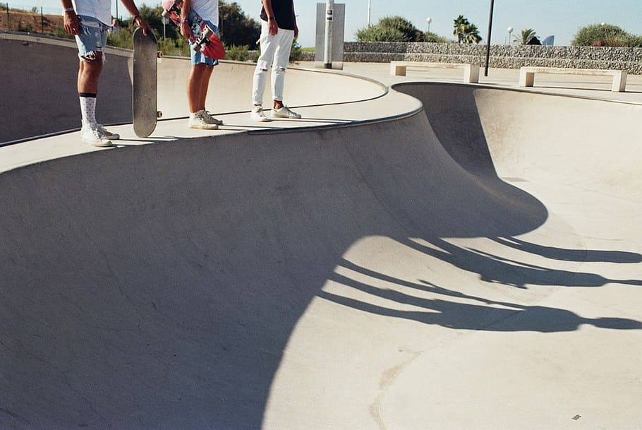 three skaters standing on skateboard concrete ramp with shadow at daytime, three persons beside gray concrete skateboard ramp