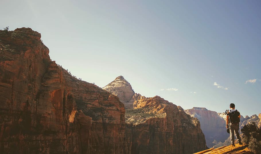 man standing on top of mountain, person standing on cliffhanger overlooking the rock formations under blue sky HD wallpaper