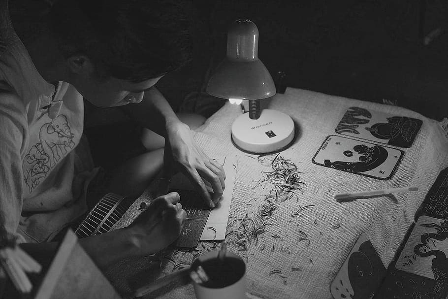 grayscale photography of man writing on brown paper near turned on lamp, person carving plate on table HD wallpaper