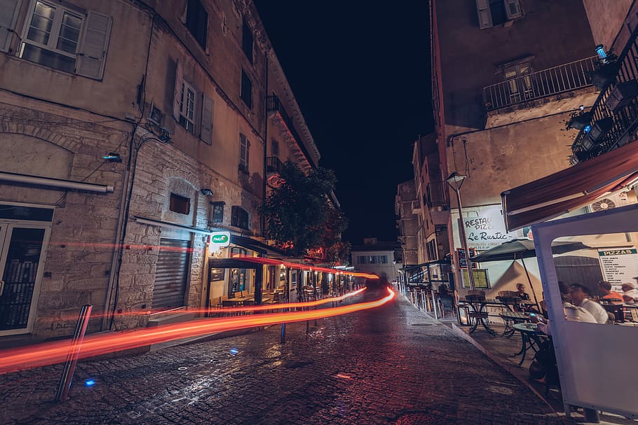 timelapse photograhy of orange light and group of people sitting on patio set beside commercial buildings during nighttime, time lapse photography of gray concrete road during nighttime HD wallpaper