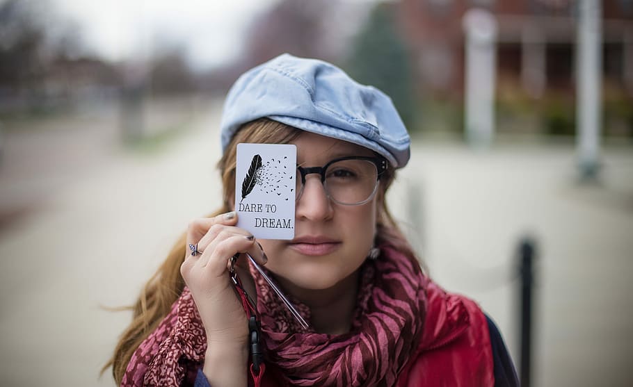 woman wearing pink and maroon zebra pattern scarf, red and black shirt, black and white plastic framed eyeglasses and blue hat while holding white Dare To Dream printed paper during daylight, HD wallpaper