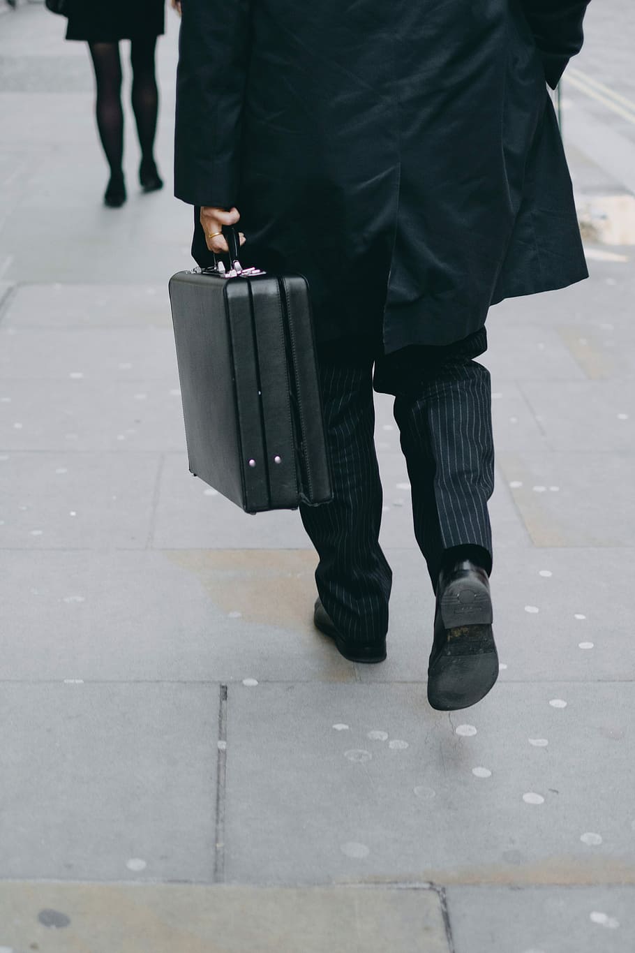 person carrying suit case while walking on pavement, man in black coat and pinstriped pants walking carrying briefcase