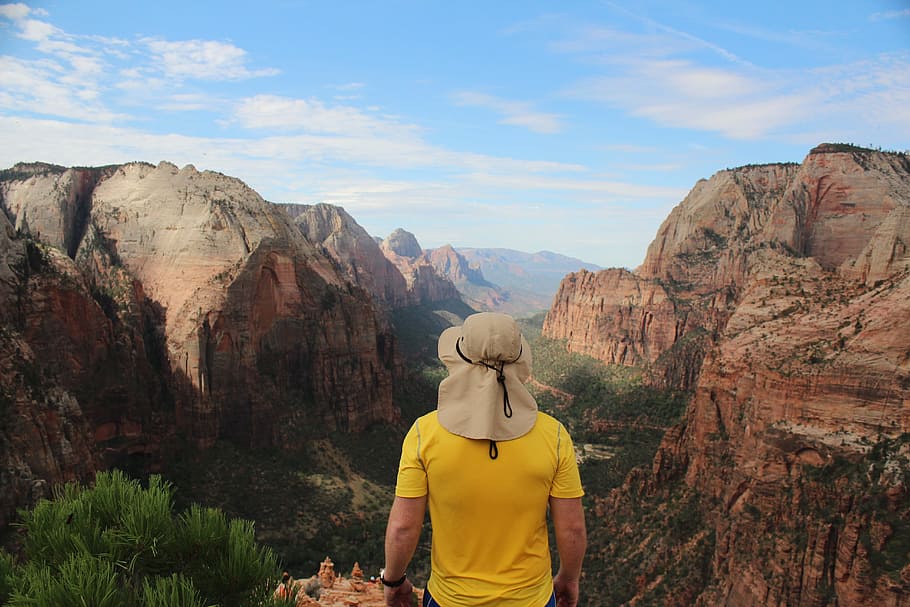 man wearing yellow shirt standing on edge of cliff facing rock formations, person in brown hat looking at rock formation during daytime