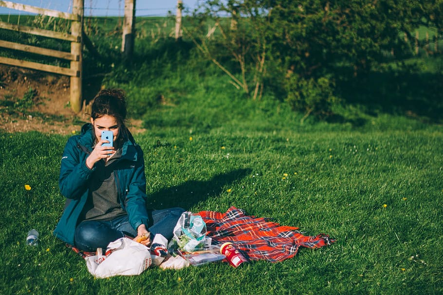 woman in blue jacket holding smartphone sitting on green grass near brown wooden fence during daytime HD wallpaper