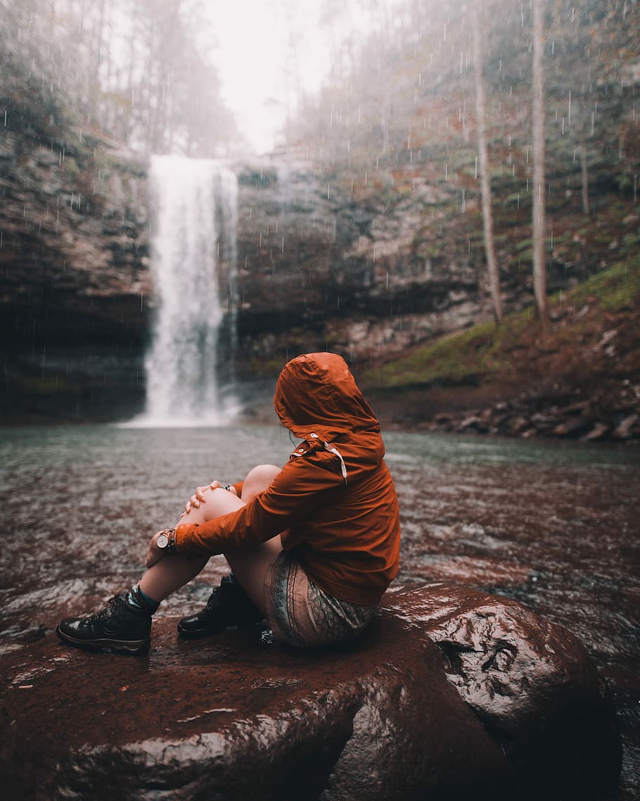 woman sitting on the rock with waterfall in distance during daytime, person wearing brown hoodie sitting near waterfalls HD wallpaper