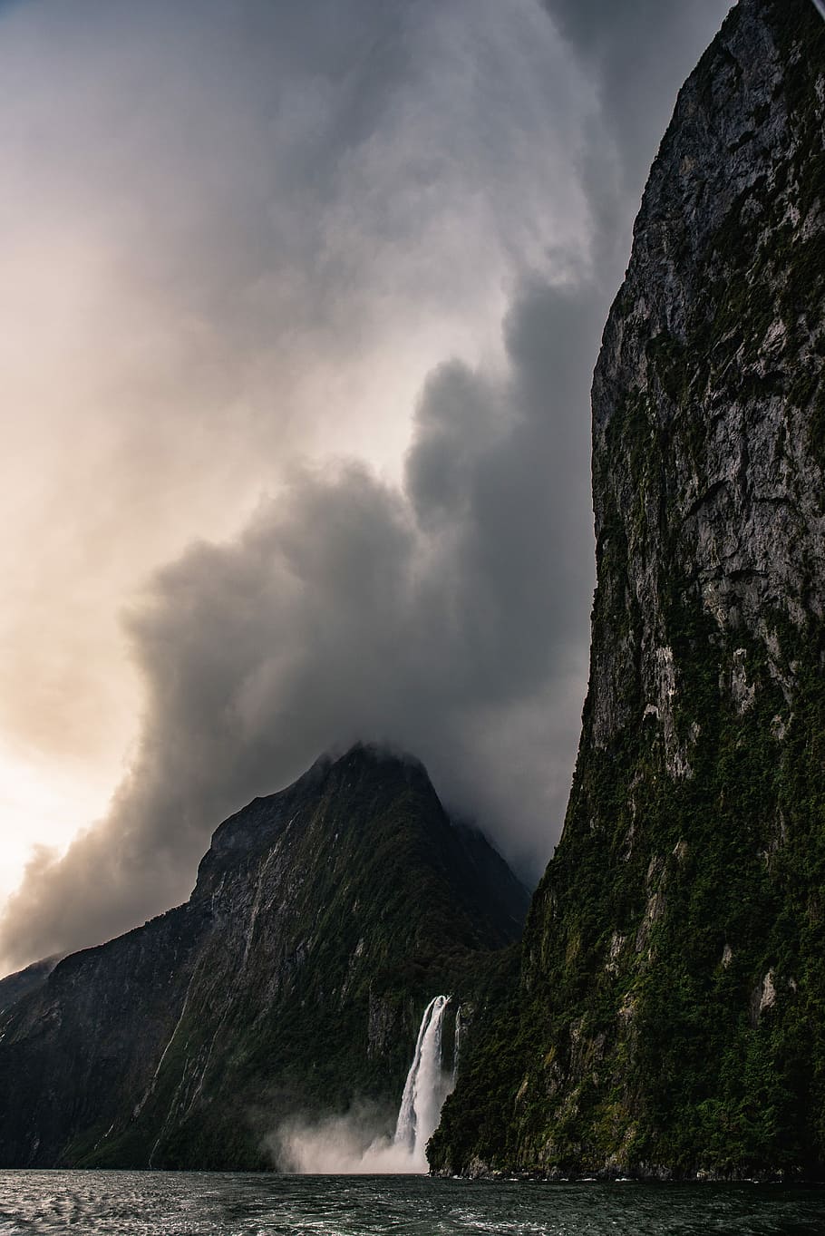 low angle photo of waterfalls between mountains, waterfalls flowing towards ocean from mountain covered with clouds HD wallpaper