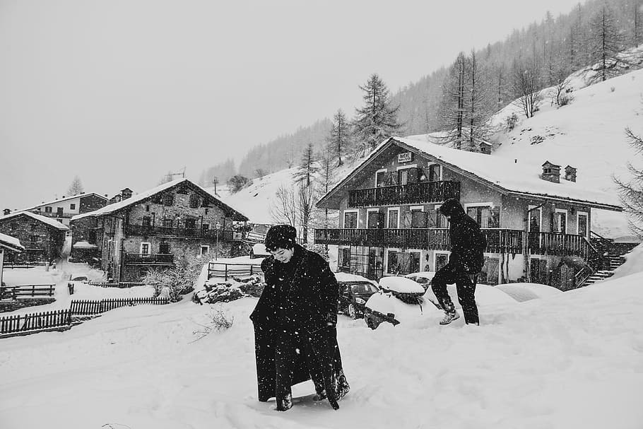 grayscale photo of man covered with coat near houses, grayscale photo of person walking on field covered with snow HD wallpaper