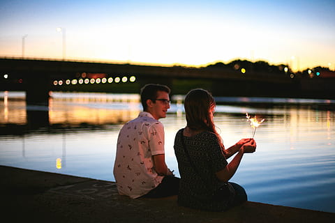 man and woman sitting beside river near bridge during sunset HD wallpaper