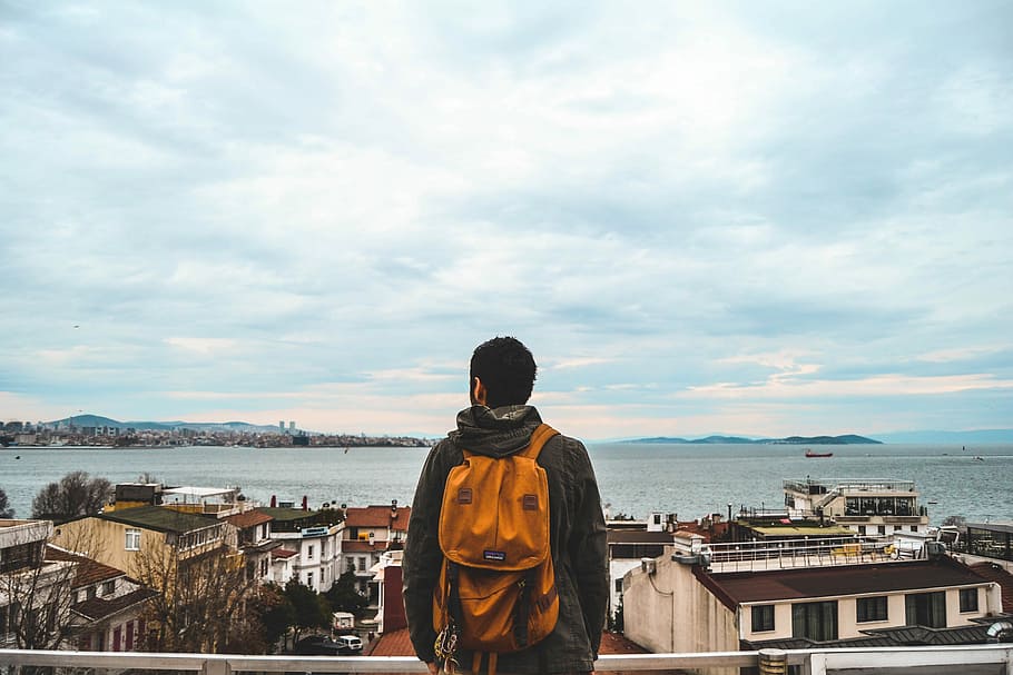 man carrying orange backpack looking at the houses near ocean, man wearing backpack and wearing hooded jacket HD wallpaper