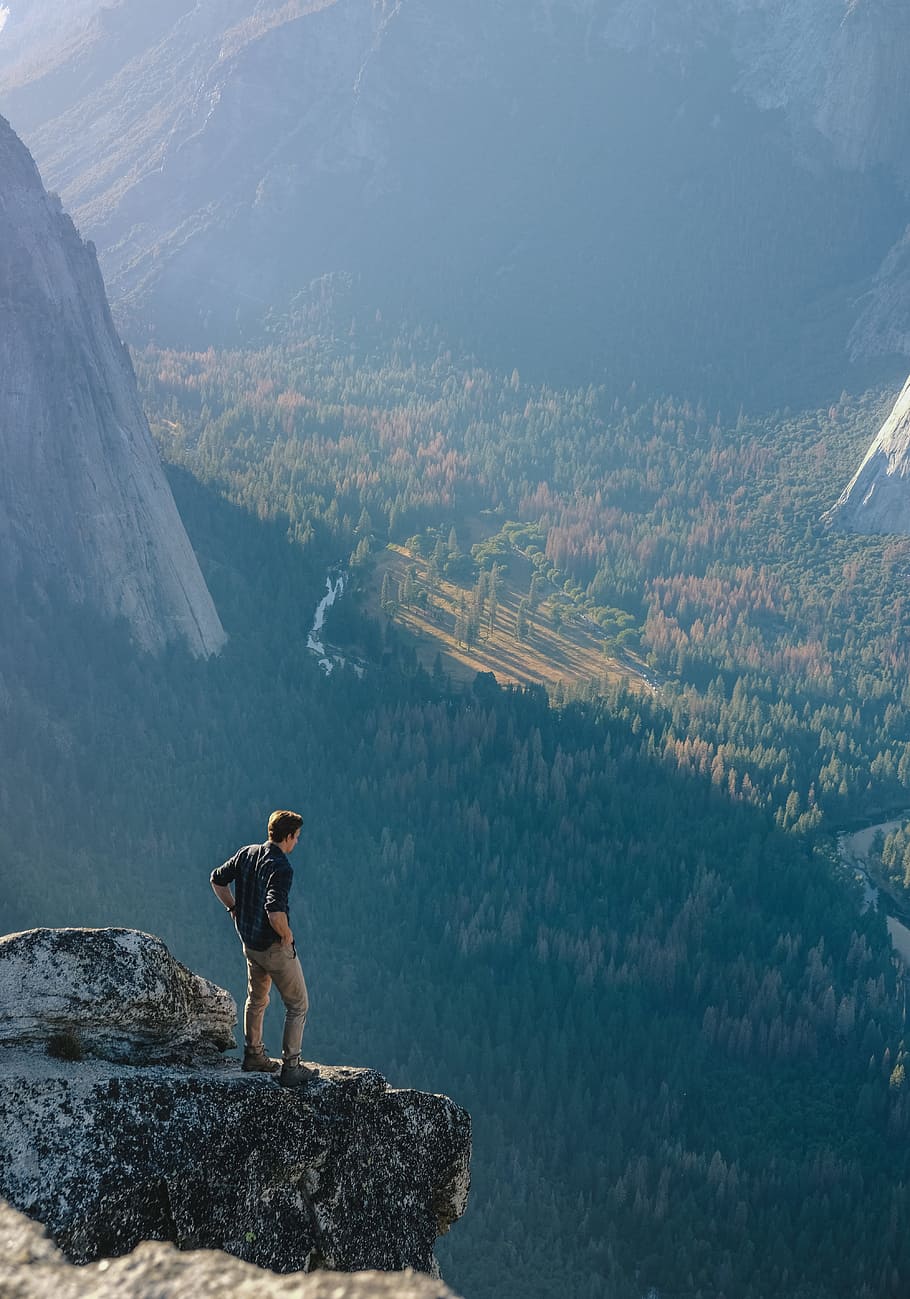 man standing on grey rock on cliff with overview of forest during daytime, man on mountain cliff during daytime HD wallpaper