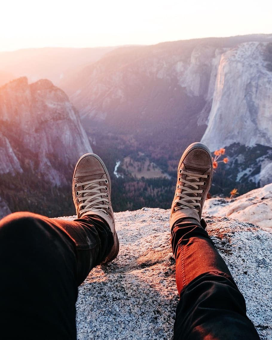 person wearing brown lace-up sneakers sitting on rocky mountain during daytime, man wearing black pant at the top of mountain during sunrise HD wallpaper