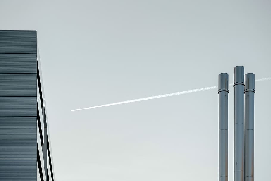 A plane with contrail is flying over an industrial building with chimneys in Geneva., three grey pipes near jet plane HD wallpaper