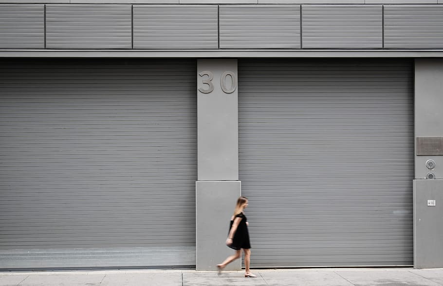woman walking beside gray shutter door, woman walking near gray metal wall, HD wallpaper