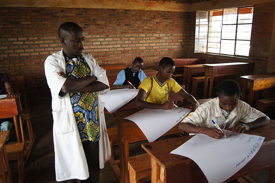 man wearing multicolored shirt and white robe outfit standing next to three men writing on paper in well-lit room during daytime HD wallpaper
