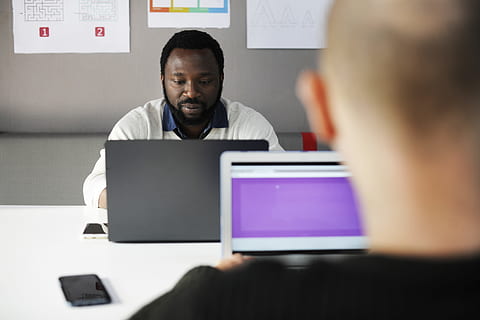 two men in front of laptop computer sitting near table with smartphone inside room, two person facing each other while using laptop computer HD wallpaper