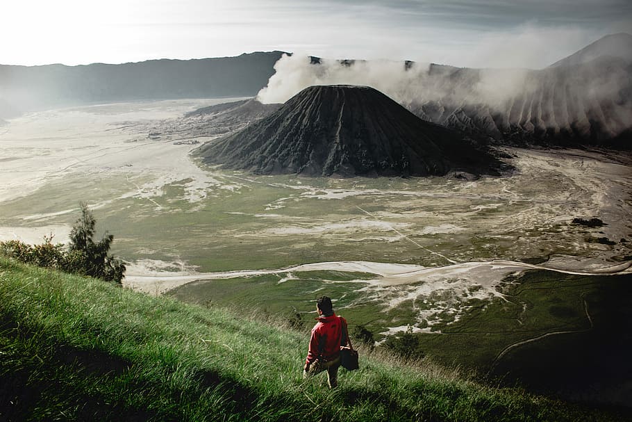 person standing on grass field near mountain, person in red jacket walking on grass near mountain at daytime HD wallpaper