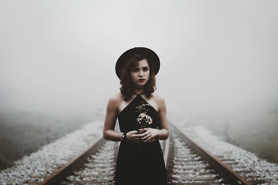 woman holding her hands on railroad, woman in black dress and hat standing in the middle of railway during daytime
