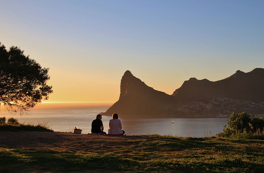man and woman sitting on green grass field watching mountain and ocean view during golden hour, HD wallpaper