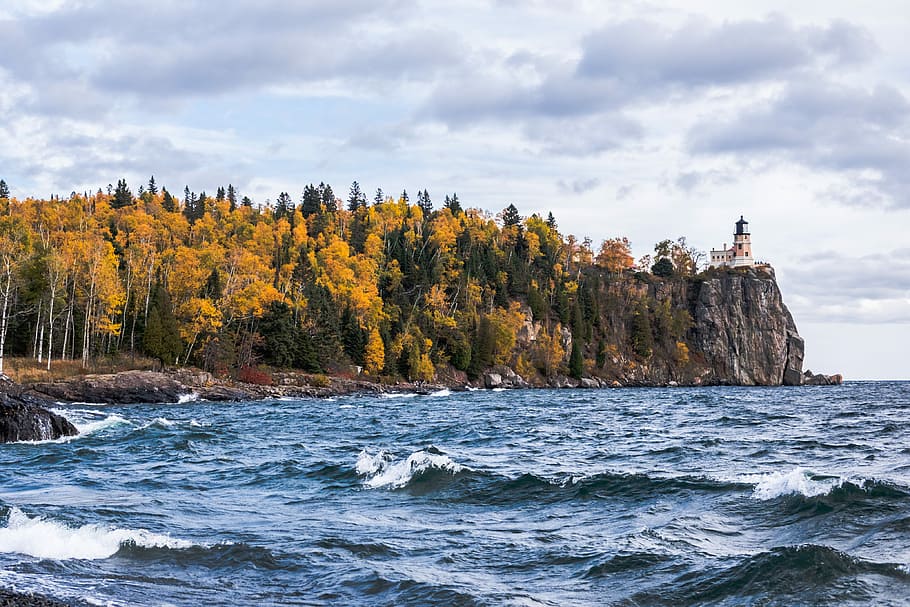 body of water near island with trees and lighthouse, lighthouse on cliff near trees and body of water at daytime HD wallpaper