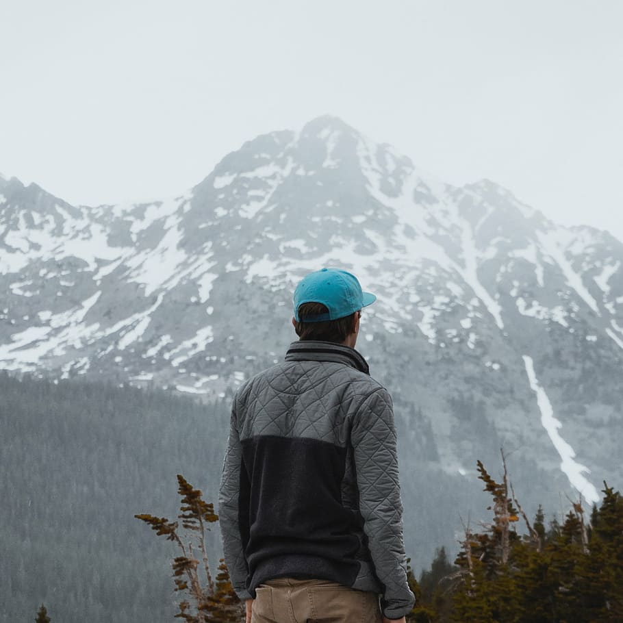 man in brown jeans wearing blue cap and gray and black jacket looking at mountain during daytime