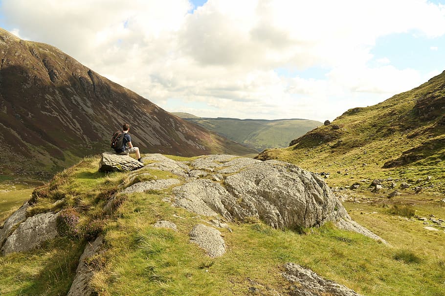 man sitting on rock near mountain during daytime, man sitting on stone near mountain under white cloudy sky during daytime HD wallpaper