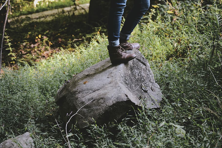 person standing on gray rock, person wearing boots standing on rock surrounded with green grass HD wallpaper
