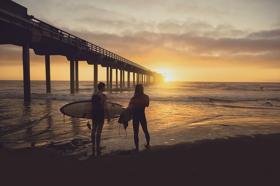 woman carrying surfboard beside person during sunset, person holding surfboard standing on seashore HD wallpaper