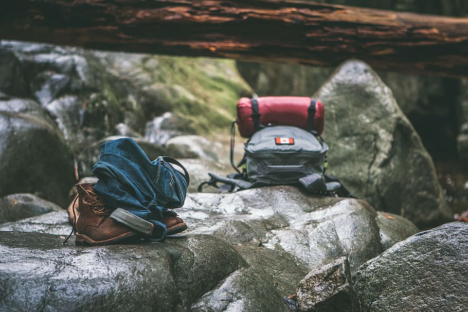 two gray and orange backpacks on gray rocks at daytime, hiking bag on rock near pair of brown leather hiking boots and blue denim jeans