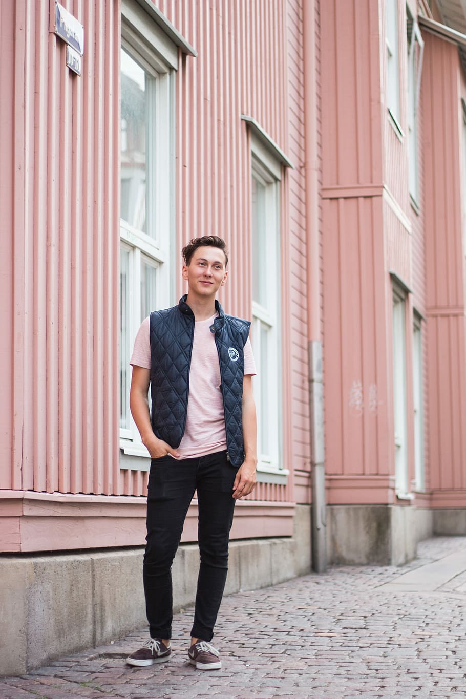 man standing beside building during day, man wearing pink shirt and quilted black vest near pink painted wall