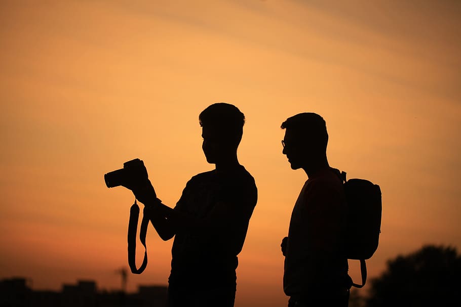 silhouette of two men carrying camera and backpack during sunset HD wallpaper