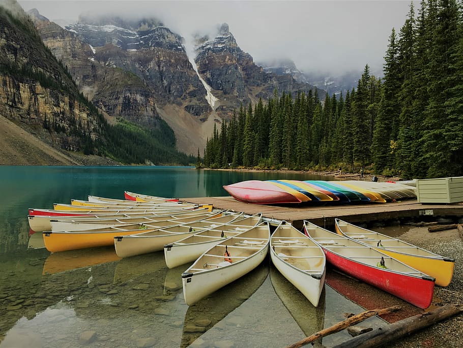 photo of assorted-color canoes on body of water surrounded by pine trees, canoes around dock near mountain ranges during daytime