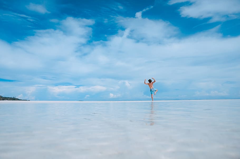 person standing in body of water, man jumping on wet sandy beach under blue cloudy skies, HD wallpaper