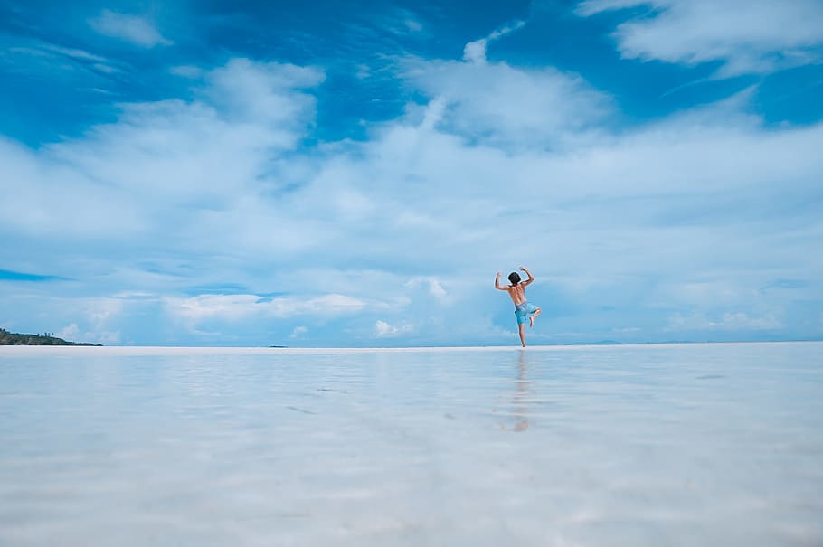 person standing in body of water, man jumping on wet sandy beach under blue cloudy skies HD wallpaper