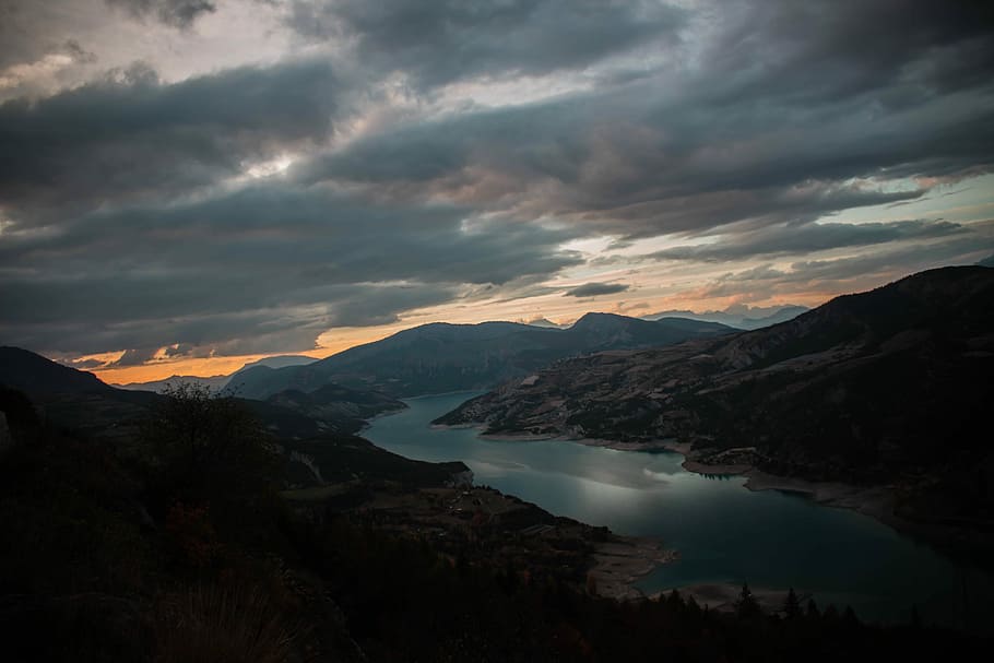 mountain beside river under gray clouds during golden hour, body of water between brown mountains under blue sky during daytime HD wallpaper