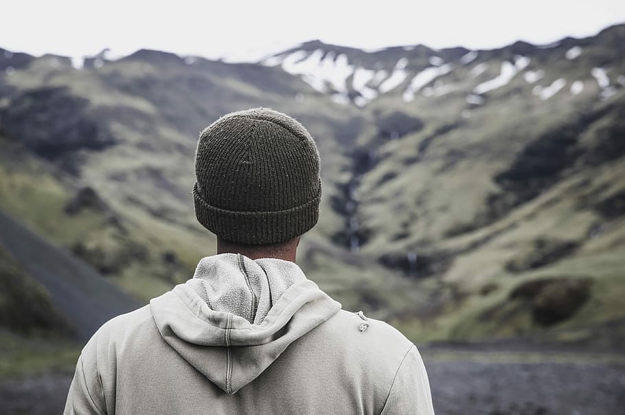man wearing gray hooded jacket in front of mountain, person in white hoodie and gray knit cap facing mountains