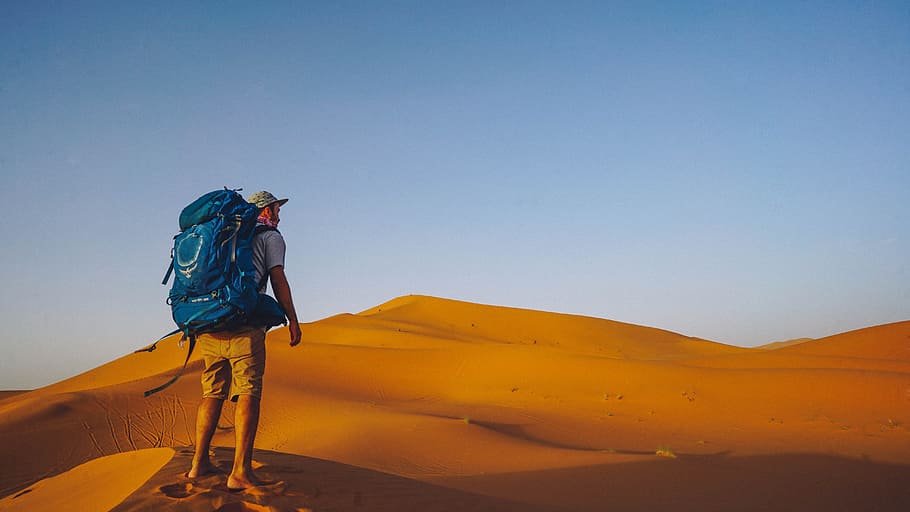 man wearing backpack standing in desert, man with blue backpack standing on dessert at daytime