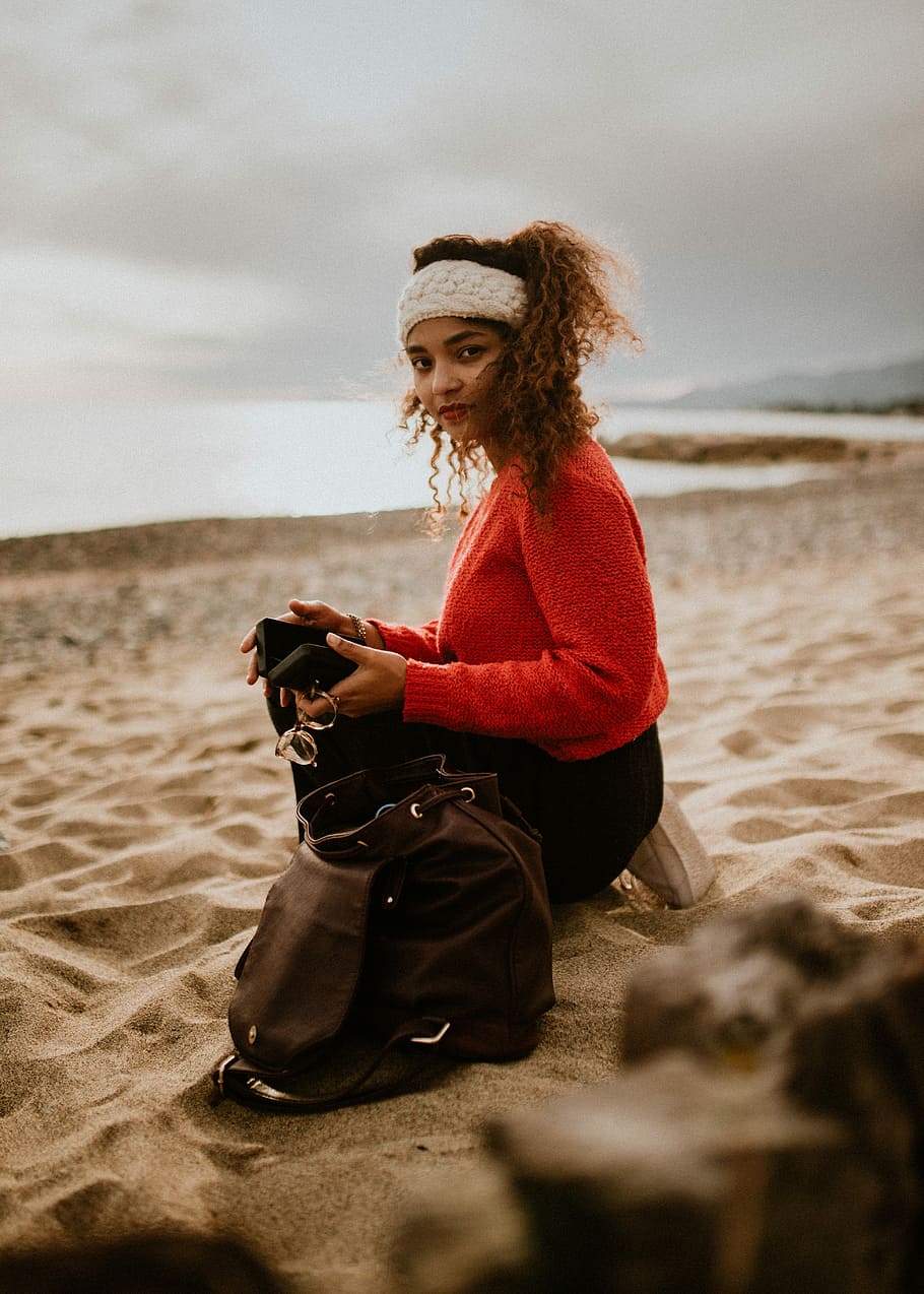 free spirit, woman holding phone while kneeling on beach sand beside her bag HD wallpaper