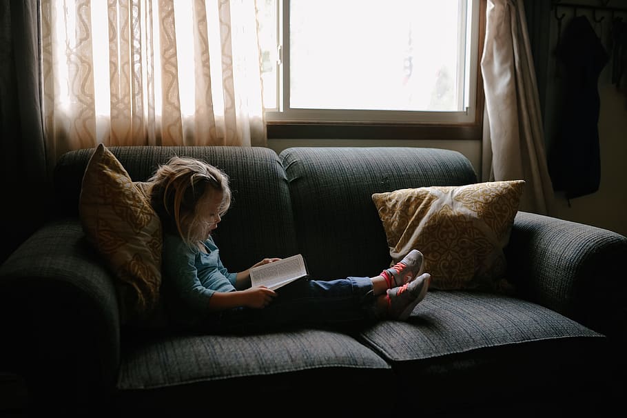 girl reading book sitting on sofa, girl sitting on sofa reading book beside closed window