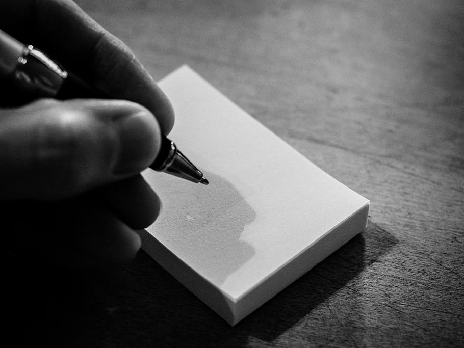 grayscale photography of person holding pen beside note wooden surface HD wallpaper