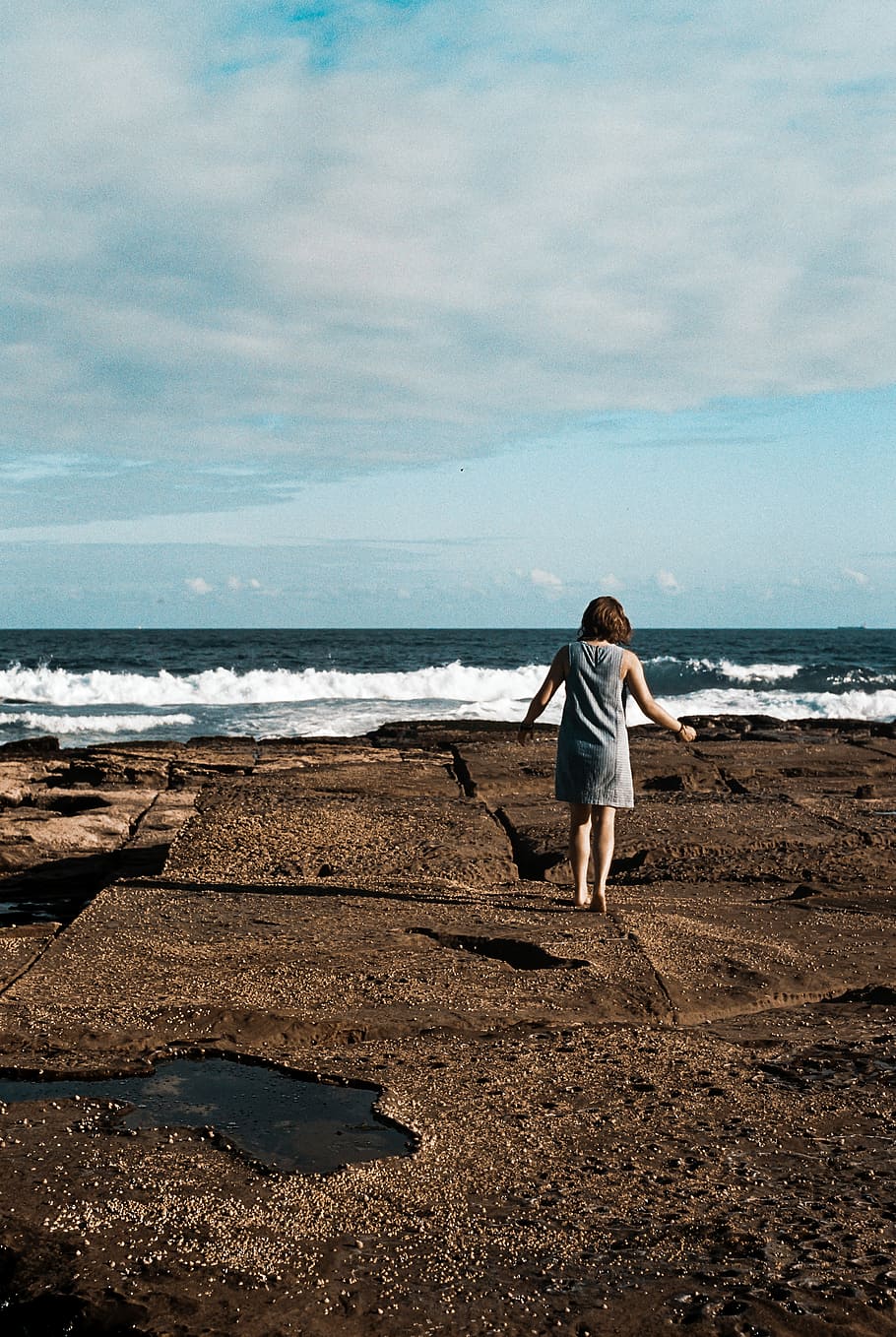 a woman walking on top of black rock formation, woman standing near ocean during daytime HD wallpaper