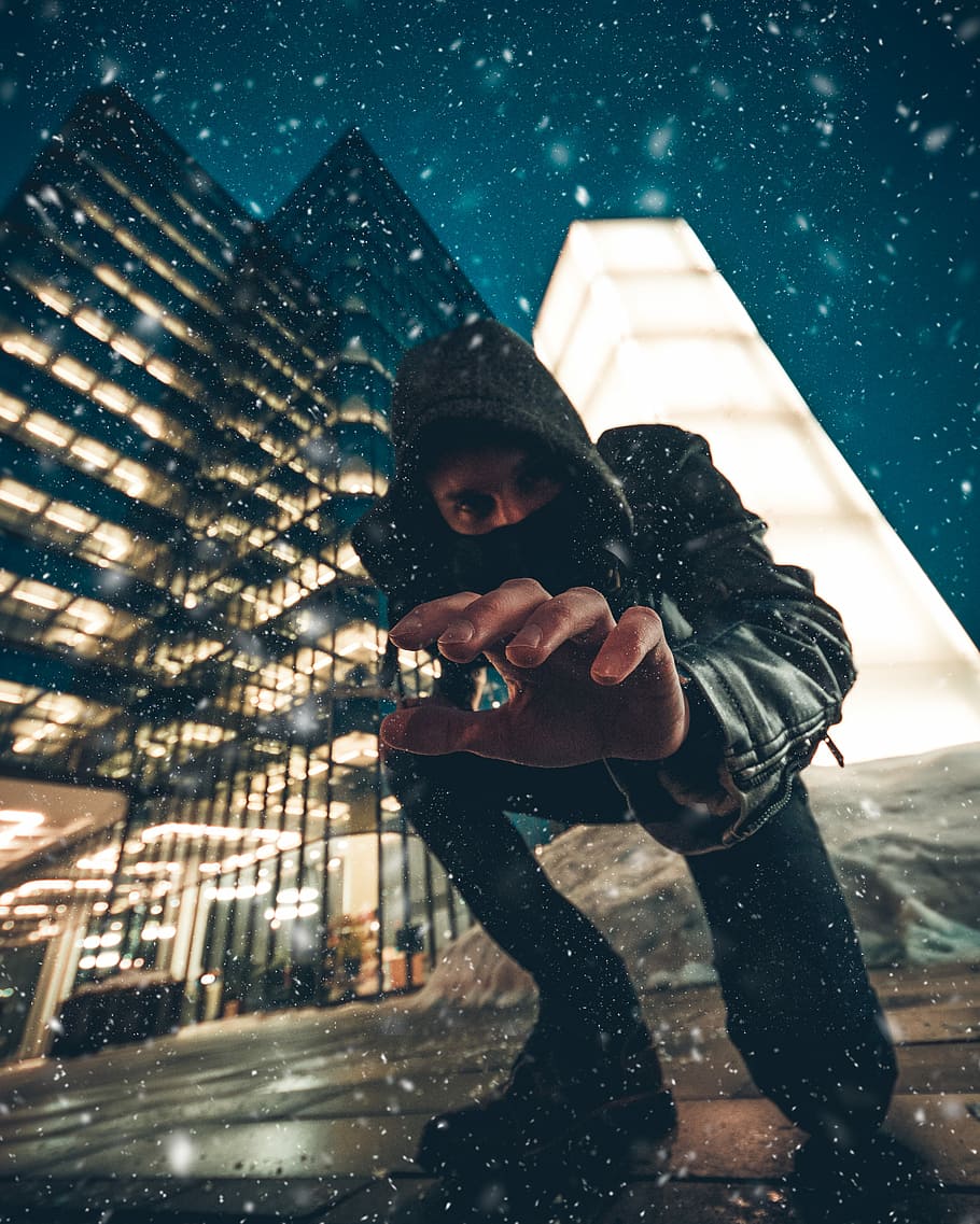 close-up photography of man kneeling on ground front of gray high-rise building during snow weather, time lapse photo of man wearing hoodie surrounded by water drops