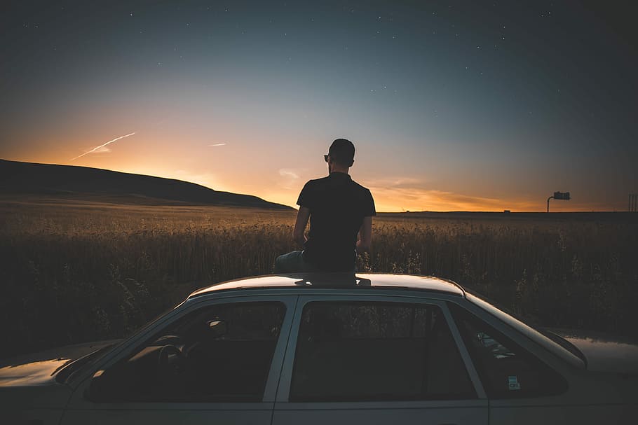 man sitting on gray sedan, man wearing black shirt sitting on top of white sedan near wheat field