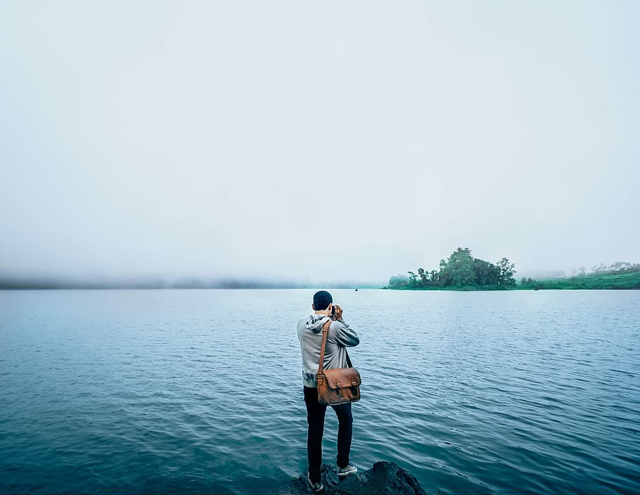 man in grey hoodie standing on rock formation near body of water during daytime HD wallpaper